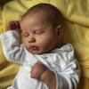 A newborn baby doll with a cloth body is lying on yellow fabric.