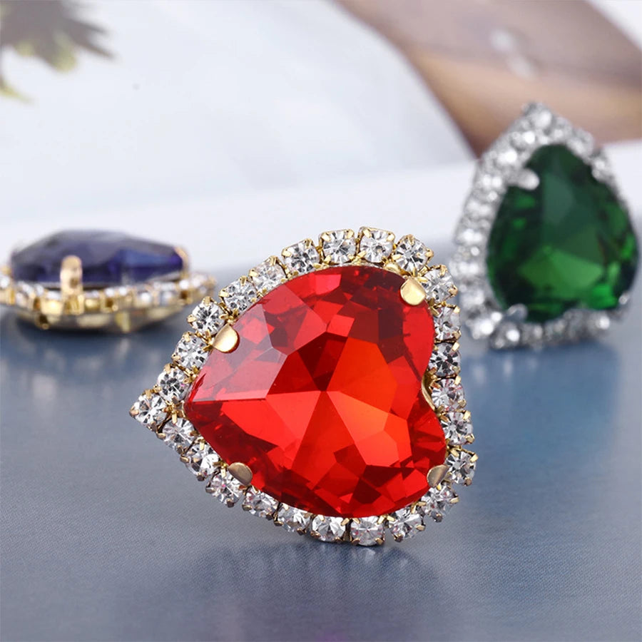 A red heart-shaped gemstone surrounded by clear rhinestones, displayed next to a green and a blue gemstone.
