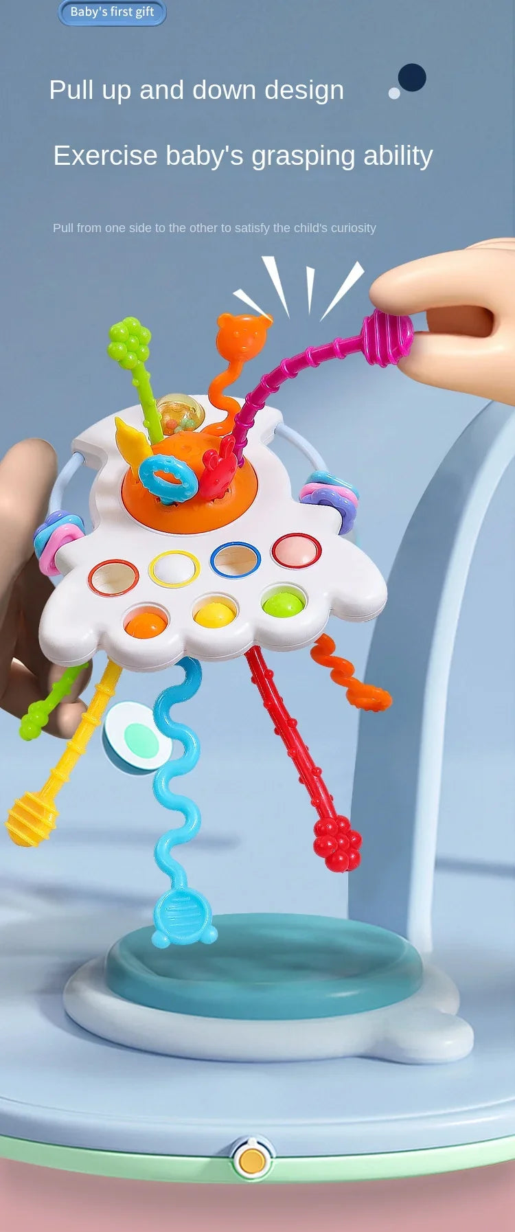 A hand is pulling a colorful string toy with various shapes and colors from a cloud-like structure for a baby's sensory exerc