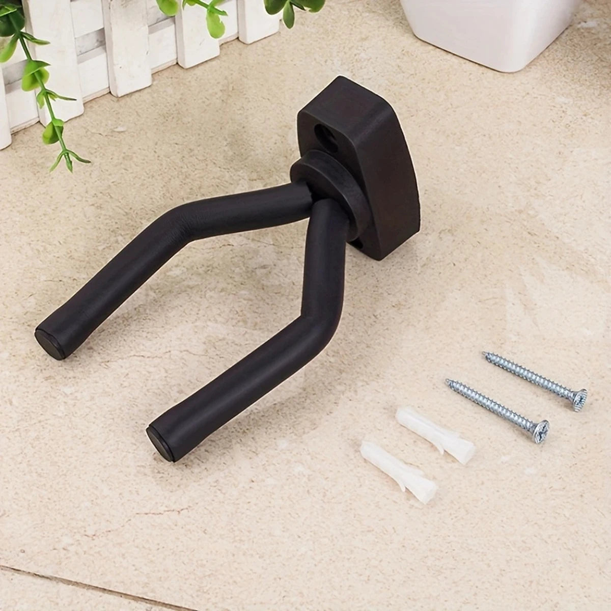 A black universal guitar wall mount hanger with threaded ends and mounting hardware lying on a light surface.