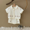 A cream-colored baby cotton shirt and shorts set with a pink flower pattern hanging on a wooden hanger against a beige wall.