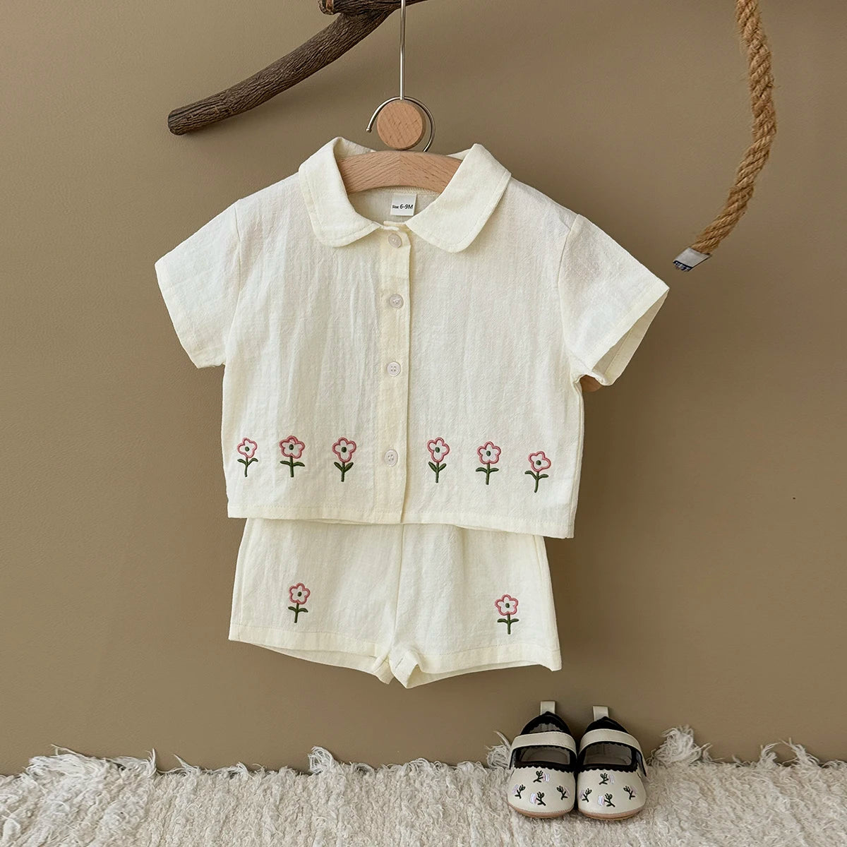 A cream-colored baby cotton shirt and shorts set with a pink flower pattern hanging on a wooden hanger against a beige wall.
