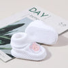 A pair of white knitted baby booties with a pink embroidered face resting on a magazine page.