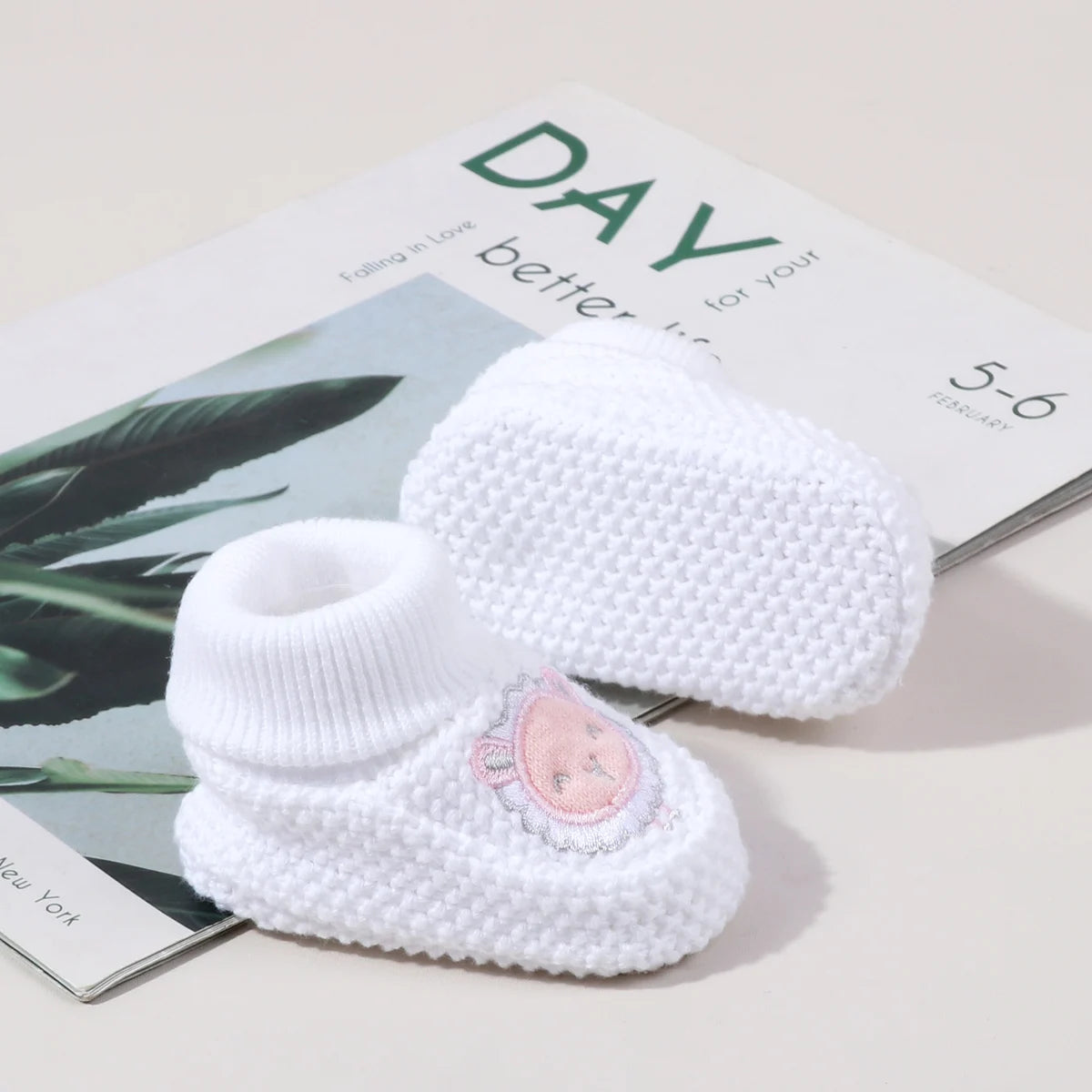 A pair of white knitted baby booties with a pink embroidered face resting on a magazine page.