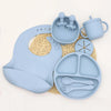 A light blue silicone tableware set for babies, including cups, plates, and utensils, resting on a woven mat.