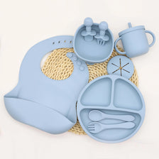 A light blue silicone tableware set for babies, including cups, plates, and utensils, resting on a woven mat.