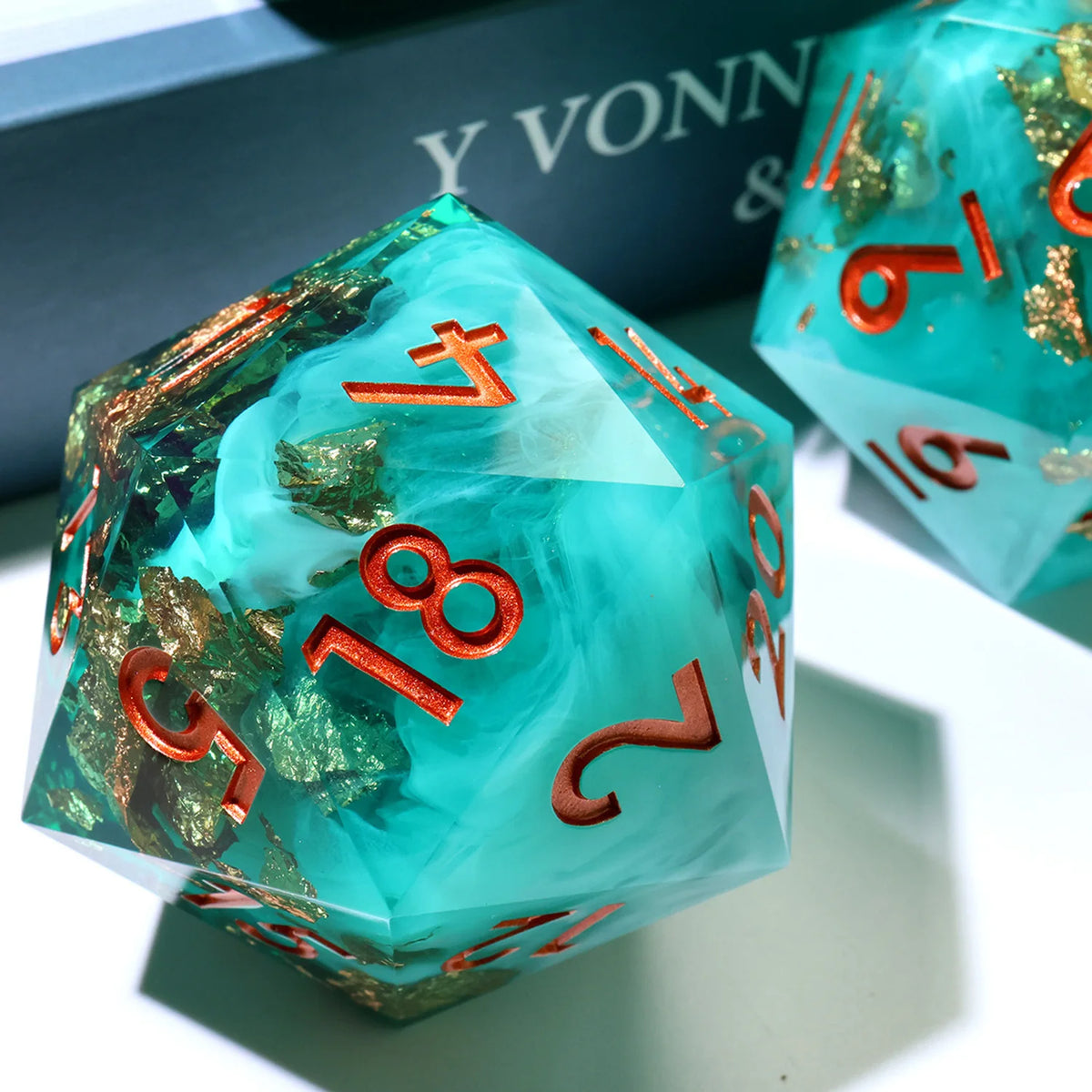 A translucent teal resin D20 die with gold flakes and orange numbers, resting on a light surface.