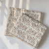 A white muslin swaddle blanket with a brown floral print is displayed under natural light.