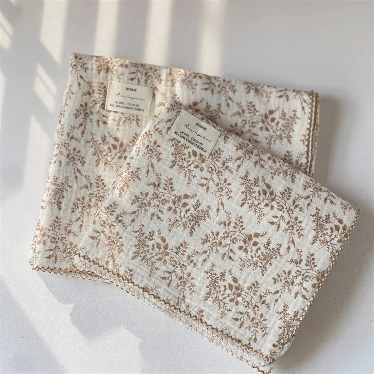 A white muslin swaddle blanket with a brown floral print is displayed under natural light.