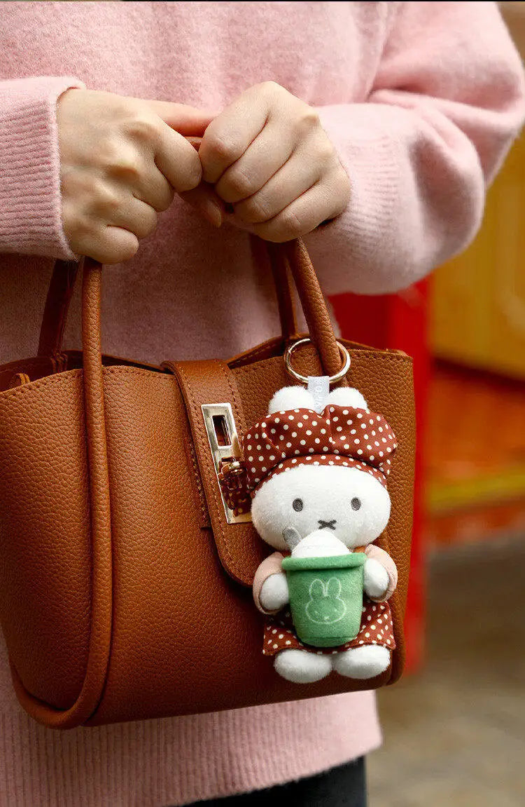 A brown leather handbag held by a person wearing a pink sweater, featuring a Miffy bakery plush keychain charm.