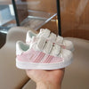 A hand holds a pair of pink and white Adidas Superstar sneakers with hook-and-loop closure.