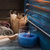 A blue candle in a glass jar sits on a wooden surface next to dried natural elements and a blue wooden background.