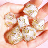 A hand holds a set of handmade resin polyhedral dice featuring gold floral designs.
