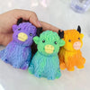 Three squishy toys shaped like Highland cows in purple, green, and orange colors