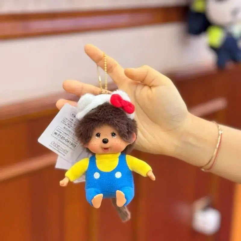 A hand holds a Monchhichi plush monkey with a red bow and yellow outfit as a charm pendant.