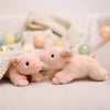 A small, pink plush pig stuffed animal lying on a white surface with soft fabric and decorative lights.