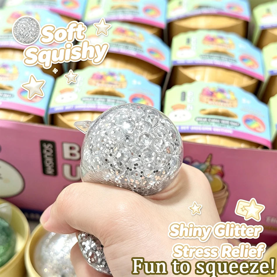 A hand holds a silver glitter ball squishy toy with a unicorn theme, demonstrating its squeezable texture.