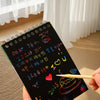 A hand holds a black scratch art book with colorful designs and a pencil against a white curtain background.
