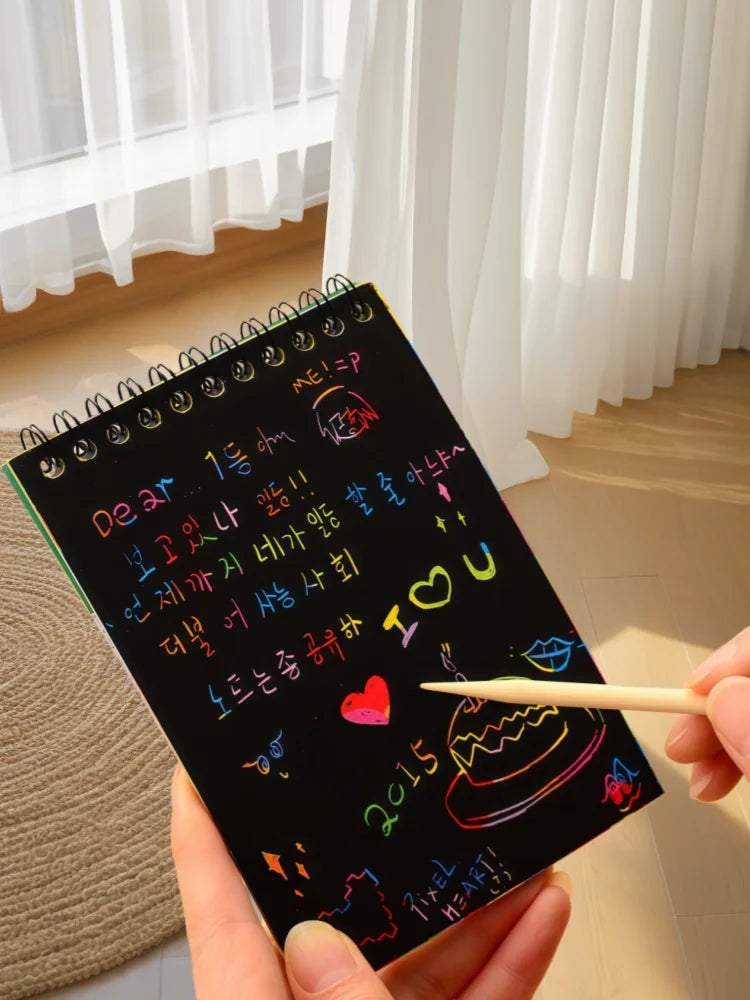 A hand holds a black scratch art book with colorful designs and a pencil against a white curtain background.