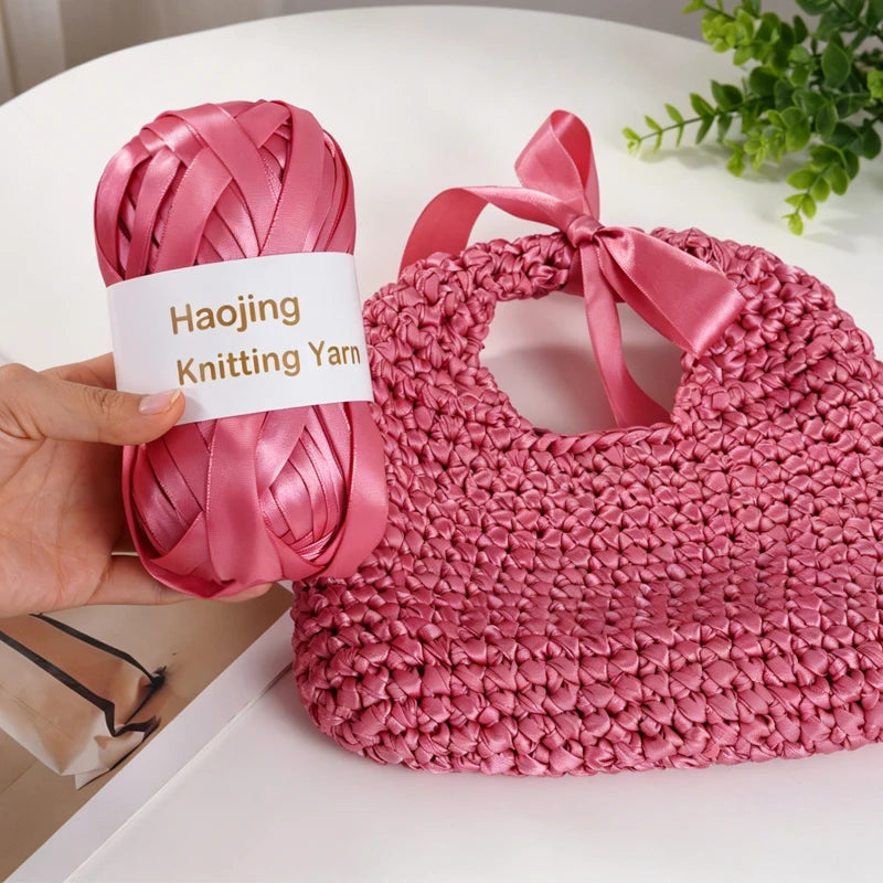 A hand holds a pink satin ribbon yarn labeled Haojing Knitting Yarn, next to a finished pink knitted bag.