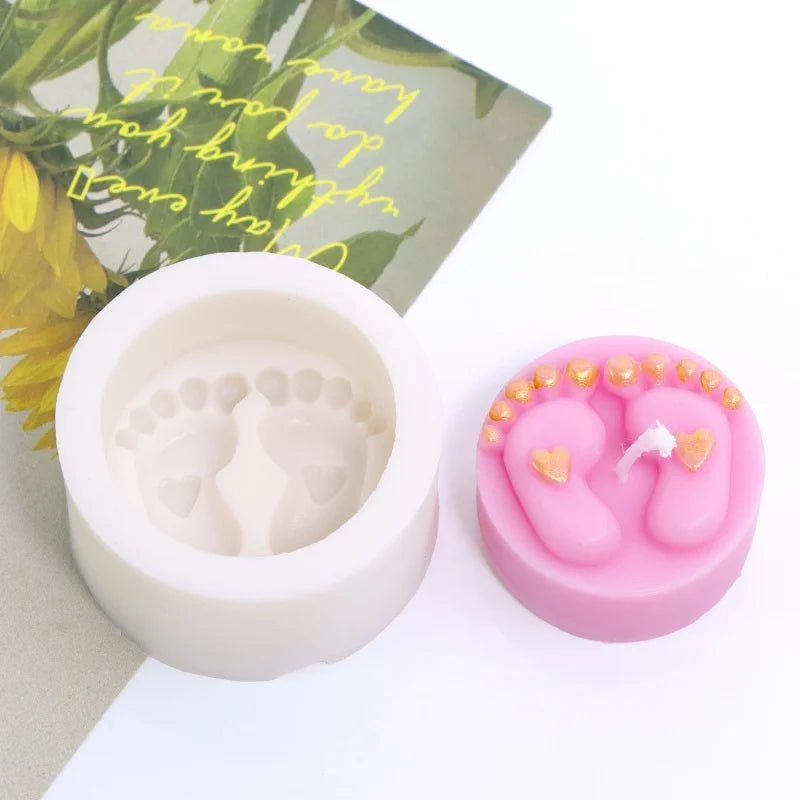A pink silicone mold shaped like a baby foot with gold hearts, next to a white mold and green leaves.