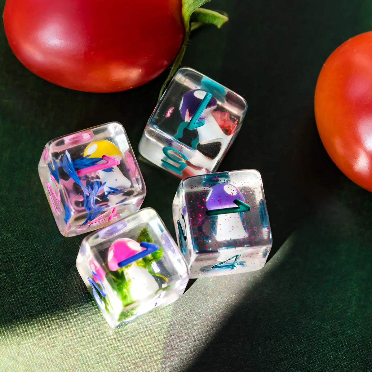 A set of translucent polyhedral dice featuring mushroom and nature themes placed on a dark green surface next to a red tomato