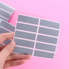 A hand holds a sheet of gray scratch-off sticker squares against a pink background.