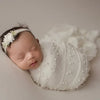 A newborn baby wearing a white knit wrap and a pearl headband, photographed in soft light.