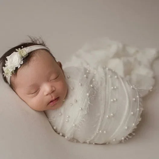 A newborn baby wearing a white knit wrap and a pearl headband, photographed in soft light.
