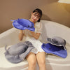 A young woman is sitting on a white sofa with two plush toy dolphins, one gray and one purple, resting on her lap.