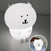A white silicone lamp shaped like a bear face with black features, illuminated on a gray surface.