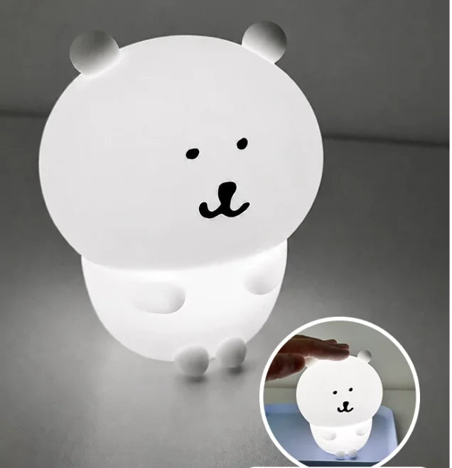 A white silicone lamp shaped like a bear face with black features, illuminated on a gray surface.