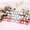 A string of silicone teething beads and clip attachments in various pastel colors and white baby name tiles.