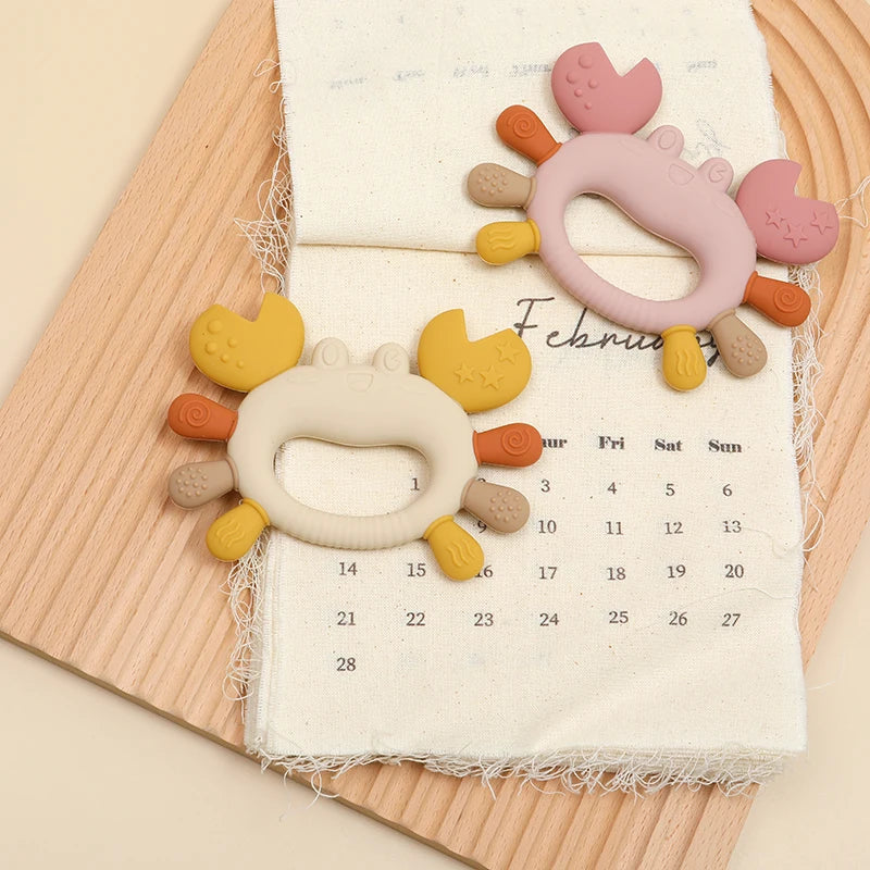 Pink and orange crab-shaped silicone baby teethers resting on a calendar page.