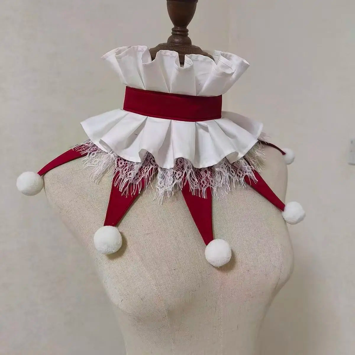 A handmade clown ruff collar featuring white fabric, red ribbon, and white pom-pom trim displayed on a mannequin.