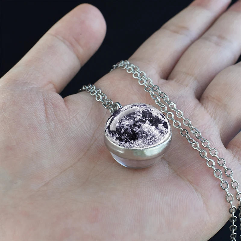 A silver pendant necklace featuring a moon-like charm with a dark, textured surface is held in a person's hand.