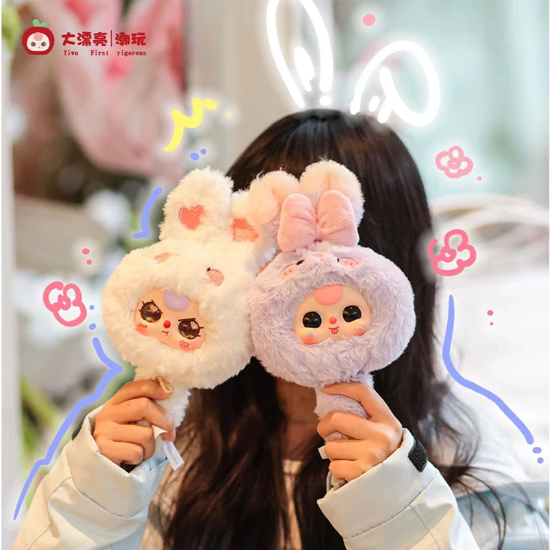 A person holds two fluffy pink and purple baby-themed collectible figures with a mirror design.