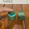 A hand holds a green fidget clicker keychain with an LED light and sound feature on a wooden surface.