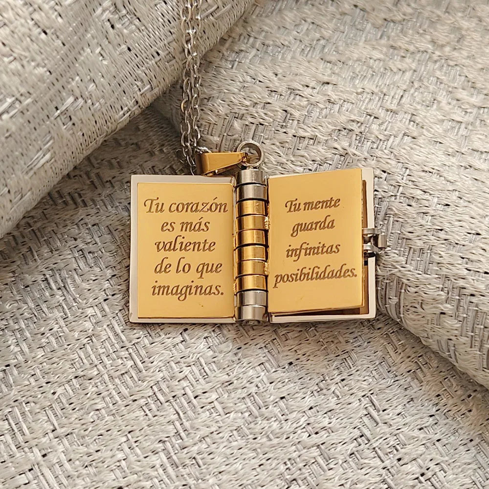 A gold-toned openable book pendant necklace featuring Spanish engraved text on a textured fabric background.
