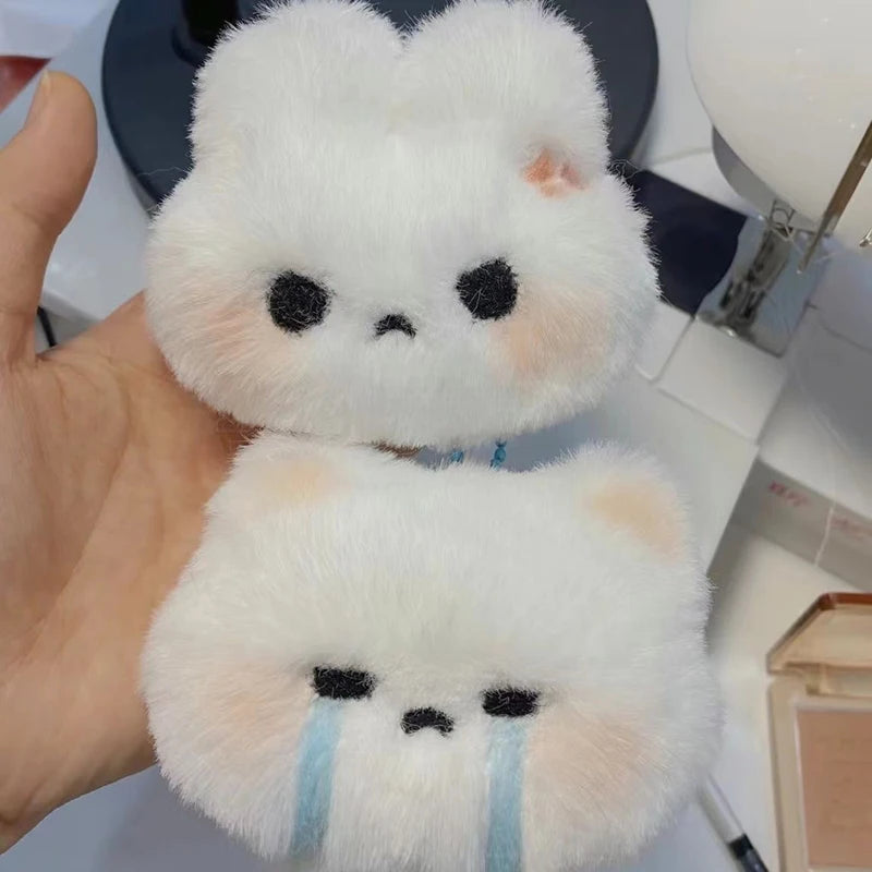 A white plush rabbit charm with black eyes and blue tear lines is held in a hand.
