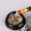 A black glass container holds small gold and silver rhinestones, with a pen tip dripping wax onto the gems.