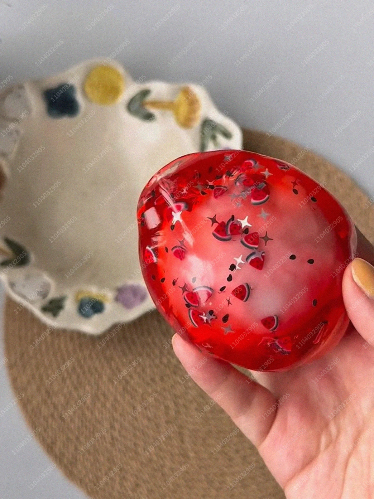 A hand holds a glossy red stress ball filled with small, shiny red and silver shapes on a brown surface.