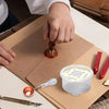 A hand is using a spoon to melt wax in a white container, set on brown paper with a heater and tool nearby.