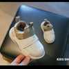 A white kids shoe with gray accents is shown resting on a dark surface.