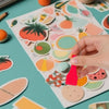 A hand holds a silver keyring attached to a red plastic lottery scratcher tool on a white card with various colorful fruit sh
