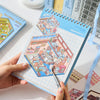 A hand holds an open sticker book featuring miniature scene decor stickers for crafting and decoration.