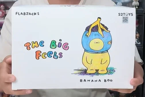 A white card featuring a colorful drawing of a yellow banana plush character with the text The Big Feels and Banana Boo.
