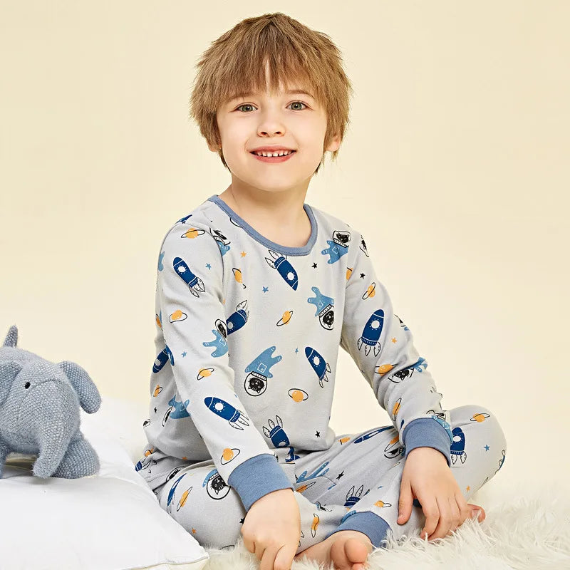 A smiling young boy wearing a light blue long-sleeved cotton pajama set with an astronaut print, sitting on a white surface.