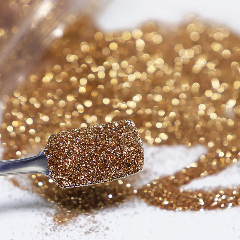 A silver spoon holds fine gold holographic glitter powder scattered on a white surface.