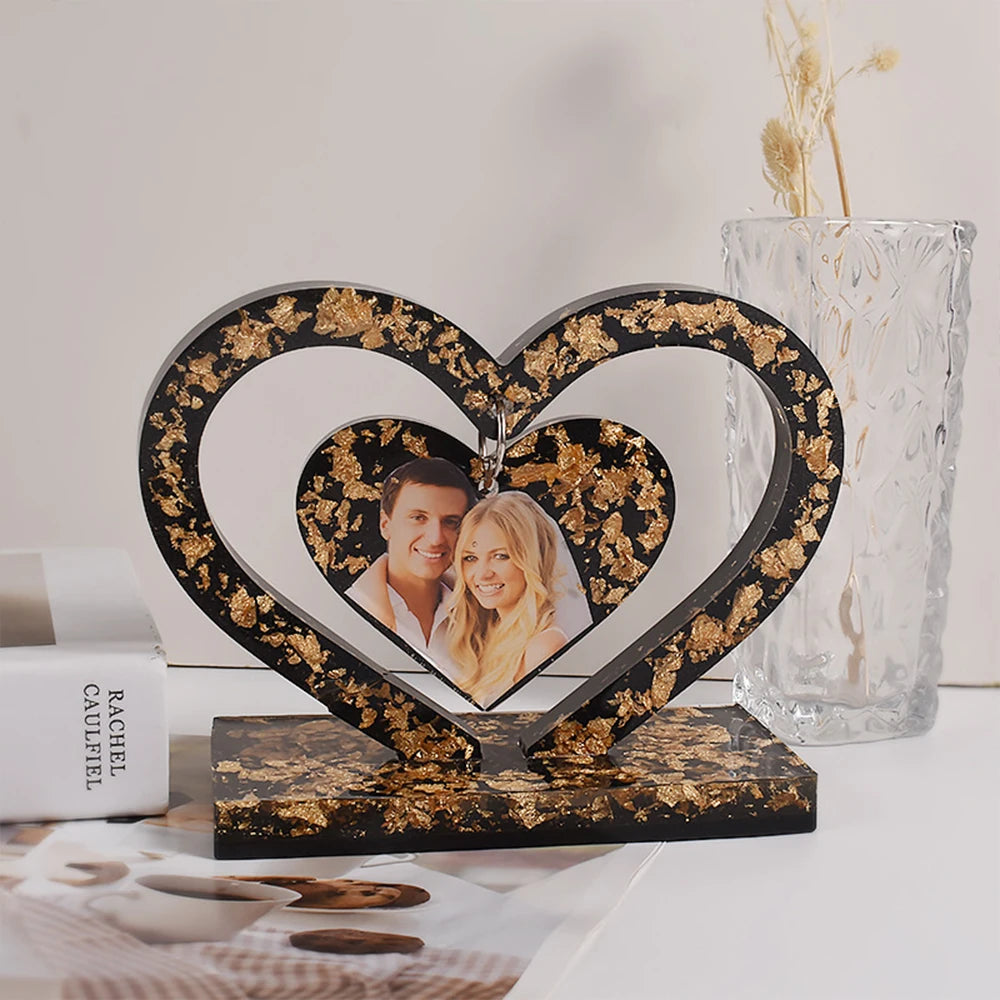 A heart-shaped frame with a black and gold floral pattern, featuring a photo of a couple, displayed on a wooden base.